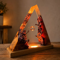 A triangular resin lamp or decorative sculpture on a wooden base, lit from within with warm amber and orange glow. The resin features swirling red, orange, and dark brown marbled patterns, with a small floating ring in the center.